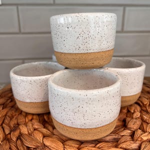 May include: Four small ceramic cups with a speckled white glaze and a natural tan base. The cups are stacked on a woven, brown surface. The cups have a simple, modern design.