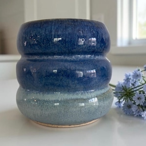 May include: A handmade ceramic vase with a unique, layered design. The vase features a gradient of blue hues, transitioning from a deep navy at the top to a lighter, seafoam green at the base. The vase has a rounded shape with a textured surface.