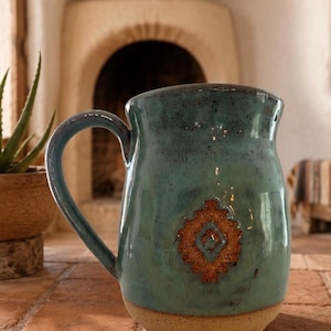 May include: A ceramic mug with a glossy teal glaze featuring dark speckles and a textured, unglazed tan base. The front of the mug displays a raised, diamond-shaped brown clay emblem with a geometric pattern centered on the body.