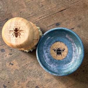 May include: A wooden dish brush and a small ceramic bowl. The brush has a round wooden handle with a bee design. The bowl is blue with a yellow center and a bee design. The items are on a wooden surface.