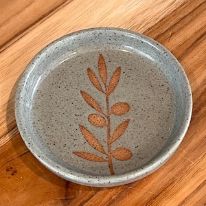 May include: A round ceramic dish with a speckled light blue-gray glaze. The center of the dish features a debossed, unglazed brown botanical design of a stem with leaves and small oval fruits, creating a textured contrast against the smooth surface.