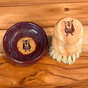 May include: A small, round, burgundy ceramic bowl and a wooden dish brush. Both items feature a black owl design. The bowl has a speckled, glazed finish. The brush has a natural wood handle and bristles.