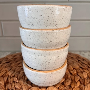 May include: A stack of four small, speckled white ceramic bowls with a natural, unglazed rim. The bowls are stacked on top of each other, showcasing their simple, yet elegant design. The bowls are sitting on a woven brown placemat.