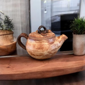 May include: A handcrafted ceramic teapot with a brown and tan speckled glaze. The teapot features a curved spout, a loop handle, and a lid with a ring handle. The teapot is a rustic, earthy design.