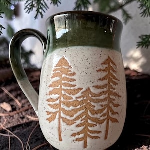 May include: A ceramic mug with a glossy dark green rim and handle, featuring a speckled off-white body. The exterior is decorated with three etched brown pine tree silhouettes that reveal the natural clay texture beneath the glaze.