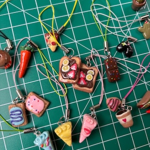 May include: A collection of miniature food charms, including toast with banana and strawberry toppings, pizza slices, and a coffee cup with a heart. Each charm is attached to a lanyard with a metal clasp.