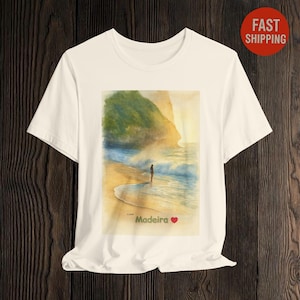 Puede incluir: Camiseta blanca con una pintura de acuarela de una escena de playa. Una persona está de pie en el agua. Las palabras "I Love Madeira" están impresas debajo, con un corazón rojo.