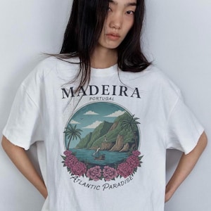 Puede incluir: Camiseta blanca con un gráfico de una escena costera de Madeira, Portugal. El diseño presenta un barco en el agua, montañas, palmeras y las palabras "Madeira Portugal" y "Atlantic Paradise" en un círculo.