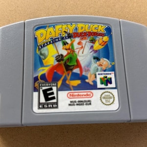 May include: A gray Nintendo 64 video game cartridge for the game Daffy Duck Starring as Duck Dodgers. The label features Daffy Duck in a green and red space suit, the game title, an E rating from the ESRB, and the Nintendo 64 logo.