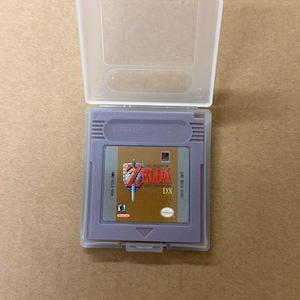 以下が含まれることがあります： 透明なプラスチックケースに入った、紫色のニンテンドーゲームボーイ用カートリッジ、ゼルダの伝説：夢をみる島DX。カートリッジには、ゲームタイトルとロゴが入った金と赤のラベルが付いています。ケースは開いています。