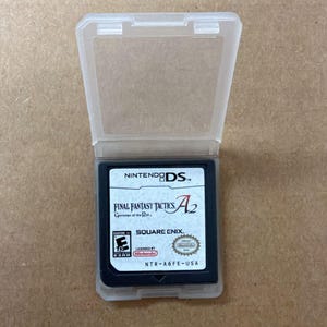 Peut inclure: Une cartouche de jeu Nintendo DS dans un étui en plastique transparent. Le jeu est "Final Fantasy Tactics A2" de Square Enix. La cartouche a une étiquette noire avec du texte blanc et rouge. L'étui est ouvert.