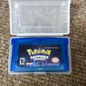 May include: A blue Pokemon Sapphire Version game cartridge in a clear plastic case. The game cartridge features the Pokemon logo and "Sapphire Version" text. The case is open, showing the game cartridge.