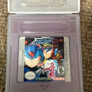 Könnte beinhalten: Eine violette Mega Man Xtreme Game Boy Color-Spielkassette in einer transparenten Kunststoffhülle. Die Spielkassette zeigt den Spieletitel und Charakterillustrationen. Die Kassette ist mit "CGB-B2XE-USA" und "CAPCOM Nintendo" beschriftet.