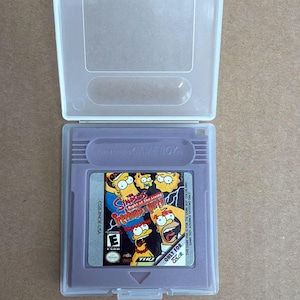 May include: A Game Boy cartridge for "The Simpsons: Night of the Living Treehouse of Horror" in a clear plastic case. The cartridge is purple with colorful graphics of the Simpsons characters. The game is rated E for Everyone.