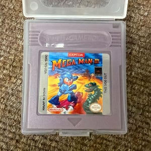 Könnte beinhalten: Eine violette Game Boy-Spielkassette für Mega Man III, mit einer farbenfrohen Illustration von Mega Man, der gegen einen grünen Drachen kämpft. Die Kassette befindet sich in einer transparenten Kunststoffhülle. Das Nintendo-Logo ist sichtbar.