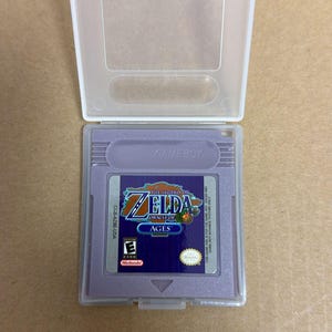 Puede incluir: Un cartucho de juego de Game Boy morado para el videojuego "The Legend of Zelda: Oracle of Ages", en una funda de plástico transparente. El cartucho tiene una etiqueta azul y dorada con el título del juego y el logotipo de Nintendo.