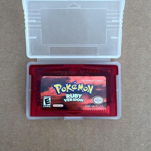 May include: A red Pokémon Ruby Version Game Boy Advance cartridge in a clear plastic case. The cartridge features the Pokémon logo and game title against a sunset background. The case is open, revealing the game cartridge.
