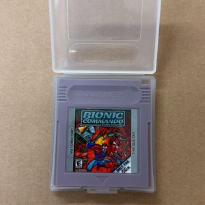 May include: A purple Nintendo Game Boy cartridge for "Bionic Commando: Elite Forces" in a clear plastic case. The cartridge has a colorful label with action figures and the text "ONLY FOR COLOR".