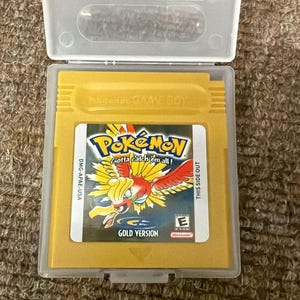 以下が含まれることがあります： 透明なプラスチックケースに入った、金色のポケモン金バージョンのゲームボーイカートリッジ。ラベルには、「ポケモン」と「Gotta catch 'em all!」の文字と、赤と黄色の鳥のような生き物が描かれています。