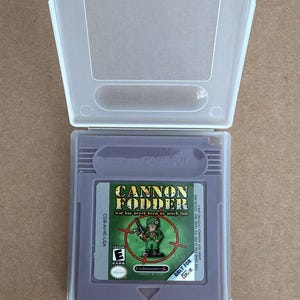 Könnte beinhalten: Eine Game Boy-Spielkassette für "Cannon Fodder" in einer transparenten Kunststoffhülle. Die Kassette ist lila mit dem Spieltitel und einer Soldaten-Grafik. Der Text "war has never been so much fun" ist ebenfalls sichtbar. Das Spiel ist "Only for Color".
