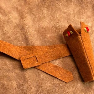 May include: A felt holster and belt set in a warm, orange-brown hue. The holster features decorative red and green accents. The belt has a rectangular buckle. The set is likely for a costume or toy.
