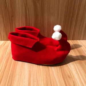 May include: A bright red elf shoe with a turned-down cuff and two white pom-poms. The shoe is made of a soft material and is designed to be worn during the holiday season. The shoe is sitting on a wooden surface.