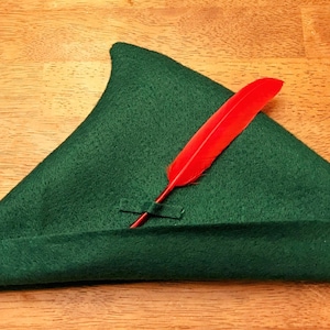 May include: A green felt hat with a triangular crown and a turned-up brim, adorned with a vibrant red feather. The hat is designed in a classic style, reminiscent of a storybook character.