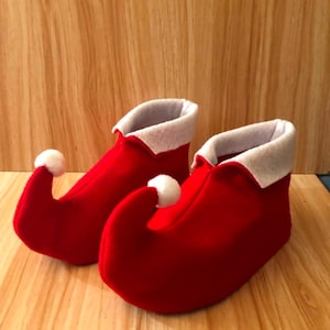 May include: A pair of bright red felt slippers featuring a curled toe design with a small white pom-pom at the tip of each toe. The slippers have a white folded cuff around the ankle opening, creating a distinct elf-inspired footwear appearance.
