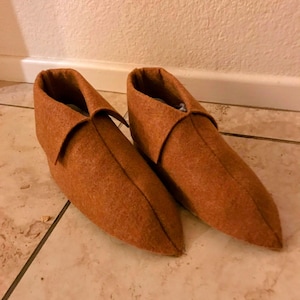 Può includere: Un paio di pantofole in feltro marrone con punta e colletto ripiegato. Le pantofole sono realizzate in un materiale morbido e strutturato e sono progettate per essere indossate in casa. Le scarpe sono appoggiate su un pavimento piastrellato.