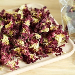May include: Dried maroon and yellow flower petals are displayed on a light wooden tray. Two clear plastic bags filled with similar petals are visible, each with a white label that reads "Flores De Rosa" in script.