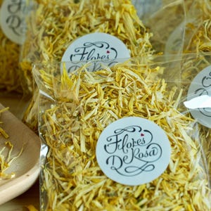 May include: Several clear plastic bags filled with dried yellow flower petals. Each bag has a white sticker with the words "Flores De Rosa" in elegant script. Some petals are on a wooden tray.