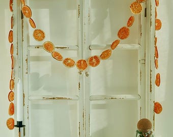 May include: A white distressed window frame is adorned with a garland of dried orange slices. A candle in a black holder, a bowl of dried flowers, and a glass jar with a cork lid are on the white surface in front of the window. The scene evokes a rustic, natural aesthetic.
