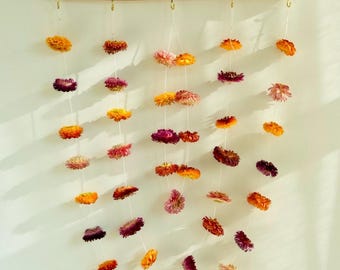 May include: A wall hanging featuring dried flowers in shades of orange, yellow, pink, and purple. The flowers are strung on white cords and suspended from a wooden dowel. The dowel is suspended by a natural fiber cord.