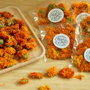 May include: Dried orange marigold flowers, some in clear plastic bags with white labels that read "Flores De Rosa". A wooden tray holds a pile of the flowers, with some loose on the wooden surface.