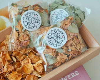 May include: A wooden tray filled with dried flower petals in shades of orange, tan, and green. Three clear plastic bags contain petals and are labeled with a white sticker that reads "Flores De Rosa" in black script.