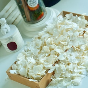 May include: A wooden tray filled with off-white flower petals. A glass jar with a cork lid contains greenery, cinnamon sticks, and a sticker with the text "Floral Day". A small white decorative container with a cherub on top is also present.