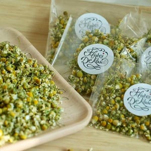 May include: Dried chamomile flowers in a wooden tray and in clear plastic bags. The bags have white circular labels with the text "Flores De Rosa" in a script font. The flowers are green and yellow.