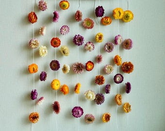 May include: A wall hanging featuring dried flowers in shades of orange, yellow, purple, and cream. The flowers are suspended from a natural wooden branch with white string, creating a decorative and natural aesthetic. The arrangement is against a light-colored wall.