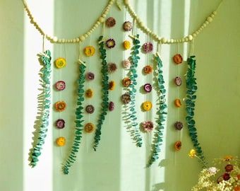 May include: A decorative wall hanging featuring dried flowers in shades of yellow, orange, and purple, interspersed with green eucalyptus leaves. The flowers and leaves are suspended from a wooden beaded garland, creating a natural and organic aesthetic.