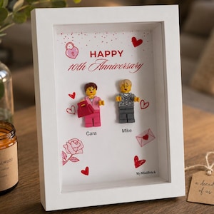 May include: A white shadow box frame containing two plastic building block figures, Cara and Mike, with red heart and rose illustrations. The text reads Happy 10th Anniversary, Cara, Mike, and My Minibrick. The background is white with red accents.