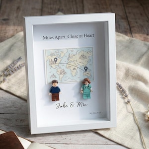 May include: A white framed art piece with the text "Miles Apart, Close at Heart" and a map illustration. Two Lego figures stand below the map, with the names "Jake & Mia" beneath them. A journal and dried flowers are in the foreground.