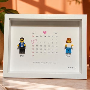 May include: A white shadow box frame containing a July 2022 calendar print with a pink heart above it. Two plastic minifigures are attached to the print, labeled Dev and Biva. The text reads, You're my always, forever yours and MyMiniBrick.