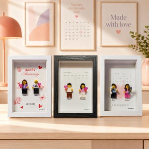 May include: Three rectangular shadow box frames containing custom minifigures and personalized text. The left frame is white with "Happy Anniversary," "Emily," "Nick," and "July 10, 2025." The center is black with a January 2026 calendar, "Zach," and "Theresa." The right is white with "Julio 2026," "Roy," "Valeria," and "te amo mucho mucho <3."