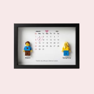 May include: A black shadow box frame containing a January 2023 calendar with a hand-drawn heart around the 9th. Two plastic toy figures stand on either side, labeled Mason and Seraphina. The text reads: You're my always, forever yours.