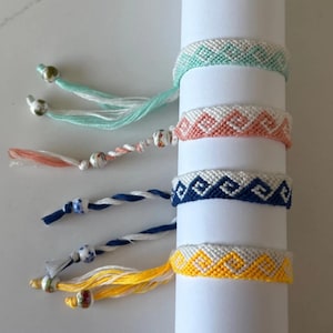 Könnte beinhalten: Eine Sammlung handgefertigter Freundschaftsarmbänder in verschiedenen Farben, darunter Mintgrün, Koralle, Marineblau und Gelb. Jedes Armband hat ein gewebtes geometrisches Muster und dekorative Quasten mit Perlen. Die Armbänder werden auf einem weißen zylindrischen Objekt präsentiert.