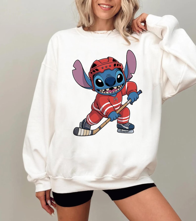 May include: White sweatshirt featuring a cartoon illustration of Stitch, the blue alien from Lilo & Stitch, dressed in a red hockey uniform and helmet, holding a hockey stick. The design is centered on the front of the sweatshirt.