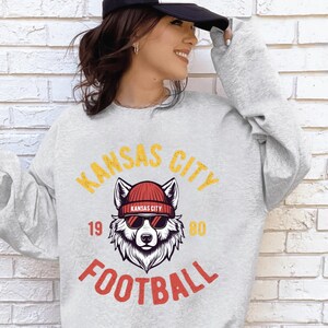 May include: A light gray sweatshirt with a graphic featuring a wolf wearing a red beanie and sunglasses. The text "KANSAS CITY FOOTBALL" is in yellow and red. The numbers "19" and "80" are also visible.