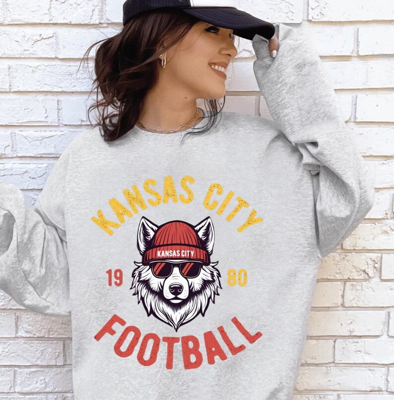 May include: A light gray sweatshirt with a graphic featuring a wolf wearing a red beanie and sunglasses. The text "KANSAS CITY FOOTBALL" is in yellow and red. The numbers "19" and "80" are also visible.