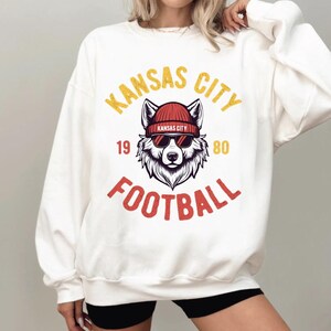May include: White sweatshirt with a graphic featuring a wolf wearing a red beanie and sunglasses. The text "KANSAS CITY FOOTBALL" is in yellow and red. The numbers "19" and "80" are also included.