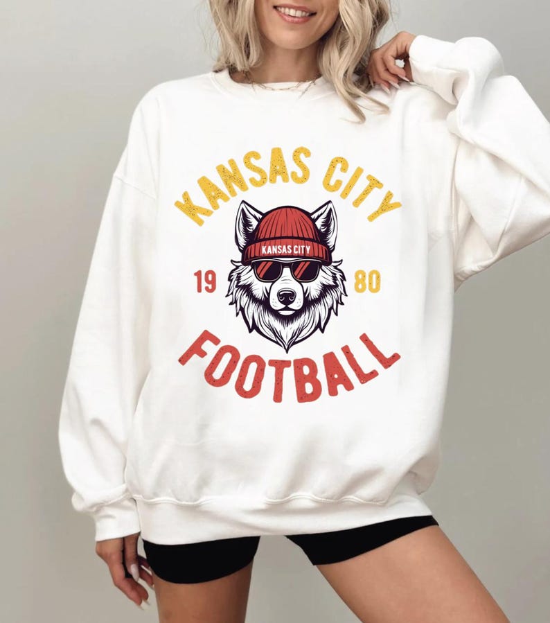 May include: White sweatshirt with a graphic featuring a wolf wearing a red beanie and sunglasses. The text "KANSAS CITY FOOTBALL" is in yellow and red. The numbers "19" and "80" are also included.
